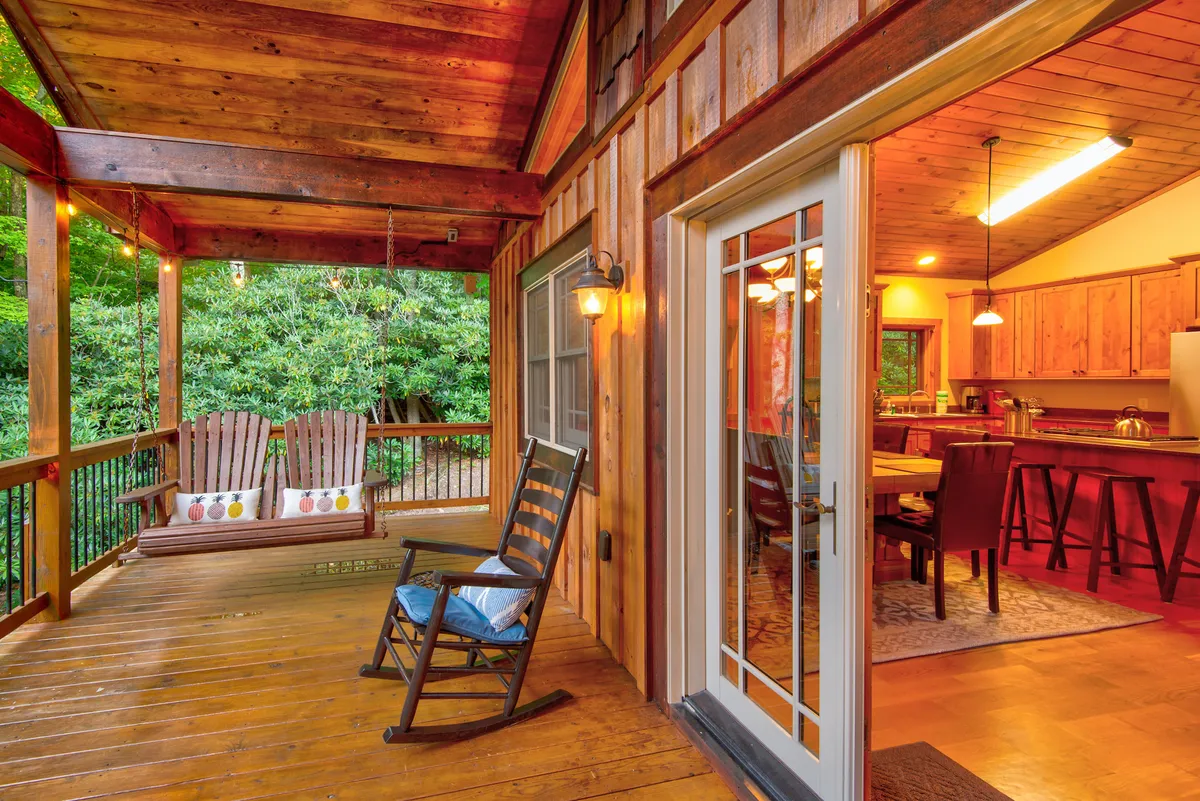 A welcoming porch scene featuring rocking chairs, swing chairs, and a cozy dining area adjacent to the kitchen entrance. The arrangement suggests both relaxation and the possibility of al fresco meals, perfect for evenings spent with friends under the stars. Warm lighting and greenery create an inviting, communal atmosphere.