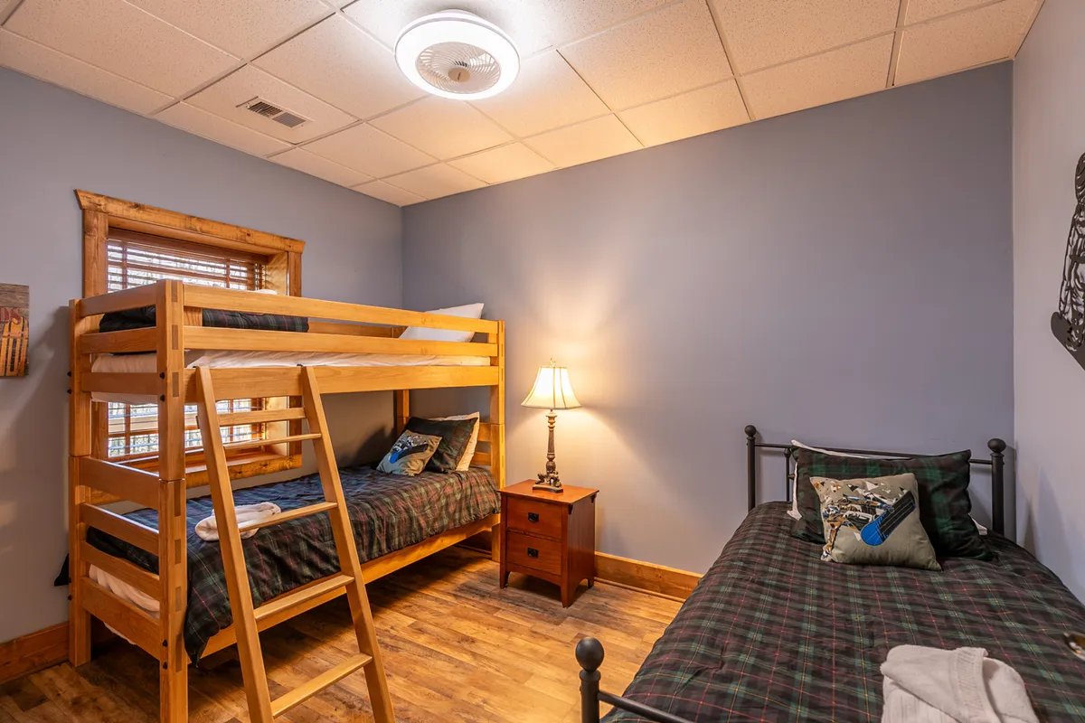 A cozy bunk room with a twin-over-twin bunk bed, a single twin bed, a nightstand with a lamp, and warm wooden furniture. The space feels playful yet restful, ideal for kids, teens, or friends enjoying a sleepover.