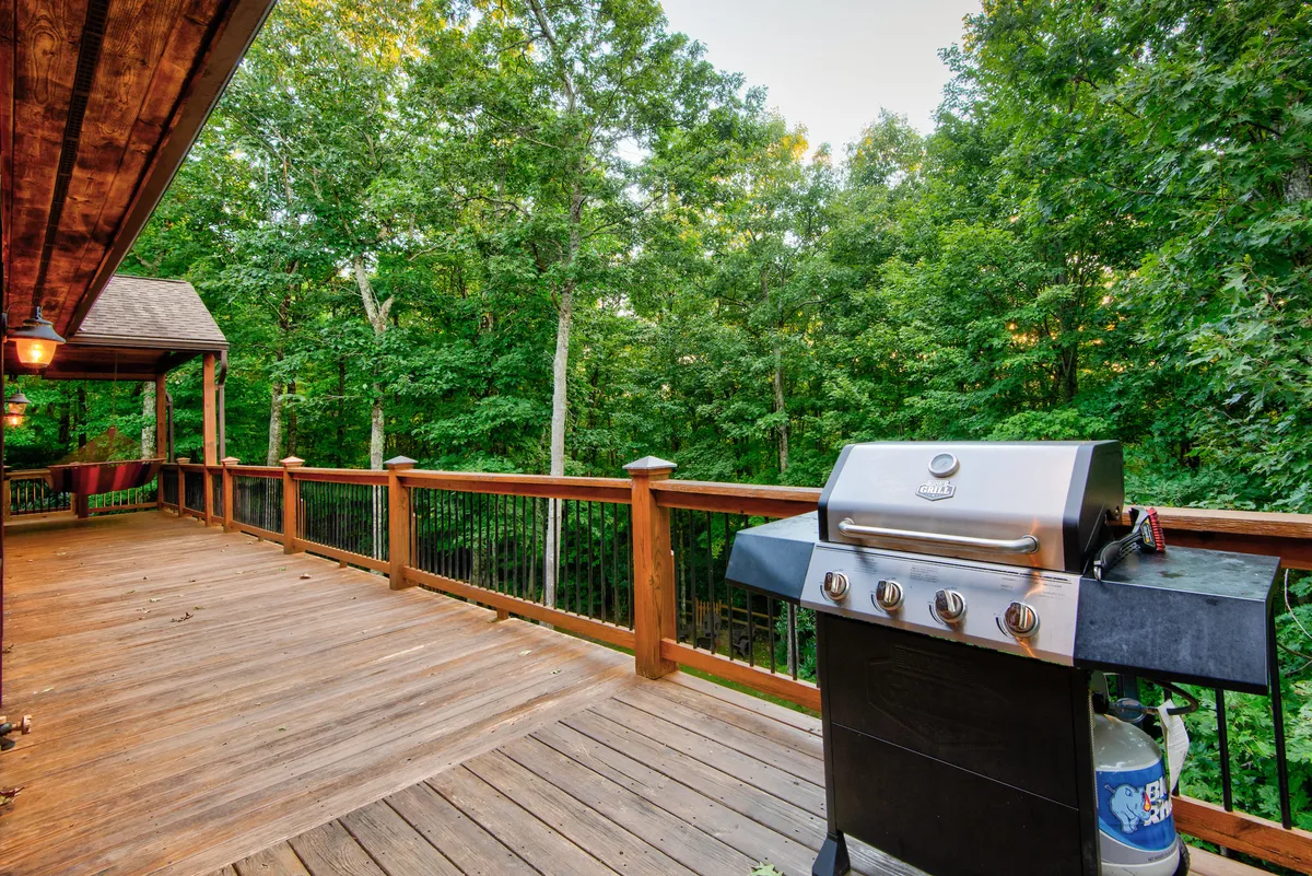 An inviting deck bordered by a wooden railing, surrounded by lush trees. The main focus is a modern grill, suggesting a perfect spot for outdoor barbecues, with ample space for gathering and relaxing. The warm, golden sunlight spills over the scene, evoking an atmosphere of late-afternoon leisure and social connection.