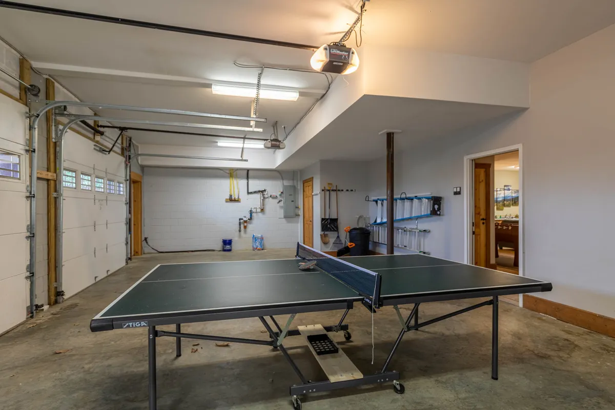 The image features a bright, open game room with a ping pong table, garage doors, and ample space for friendly competitions. Storage shelves add a practical touch, making the area suitable for families to unwind or play together after outdoor activities.