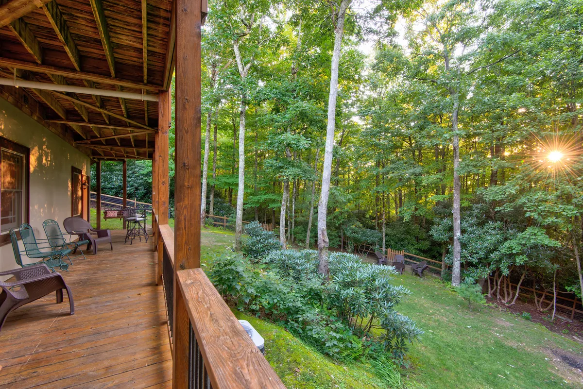 This image shows a welcoming wooden porch surrounded by lush green foliage, with sunlight filtering through the trees. The porch extends into a spacious backyard area, ideal for relaxing outdoors, hosting gatherings, or letting pets roam freely.