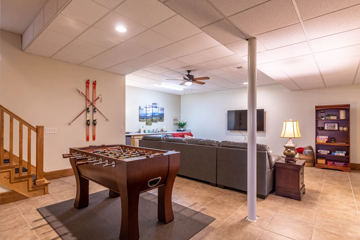 A lively game room featuring a foosball table in the foreground, a comfortable sofa seating area, a wall-mounted TV, decorative wall art, and a visible staircase leading up. The space is brightly lit, with playful vibes and modern touches.