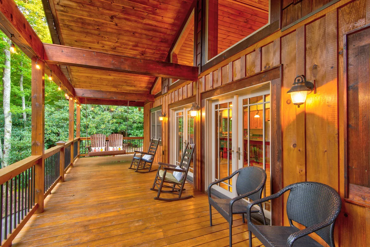 This photo showcases several rocking chairs arranged on a wooden deck, surrounded by verdant foliage. Warming hanging lights add ambiance, and the expansive outdoor setting invites quiet moments spent gazing at sunsets, chatting, or sipping coffee.