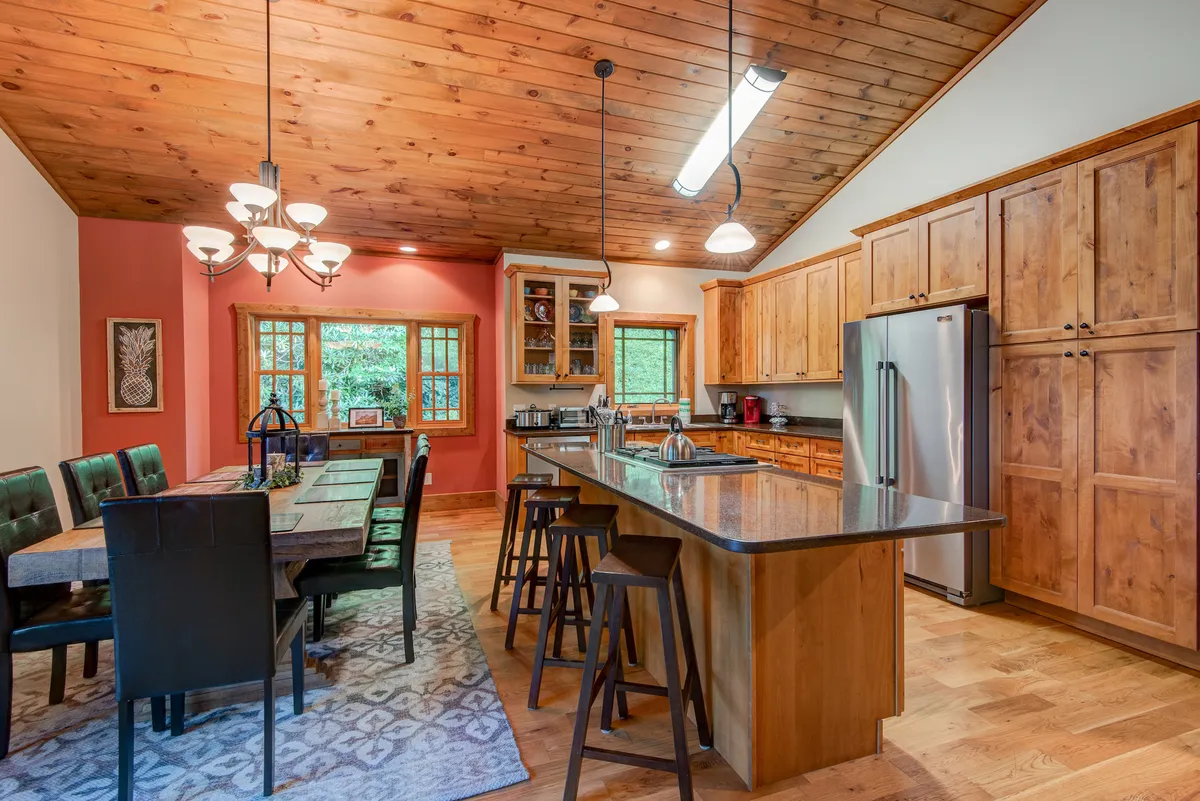 A sun-drenched dining and kitchen area featuring a spacious table, large kitchen island, wooden cabinetry, and wide windows that frame lush mountain scenery. The space is set for lively family meals and collaborative cooking, emphasizing togetherness and function.