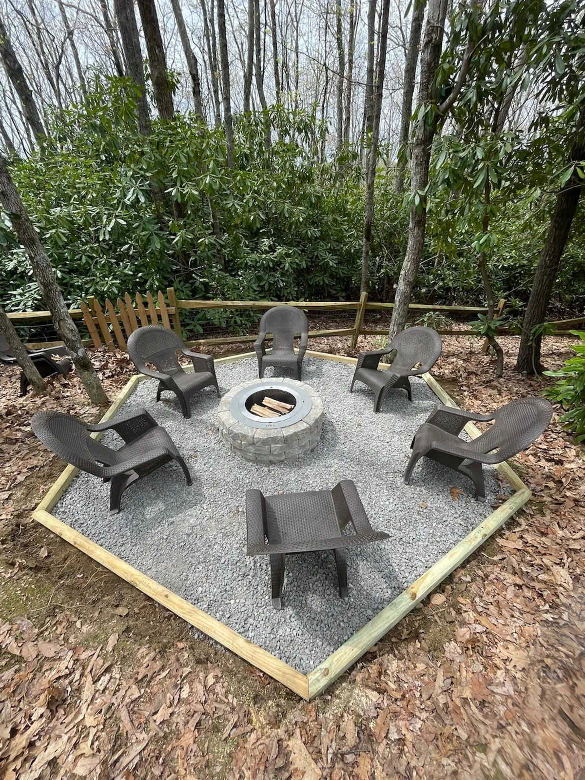 Nestled in the backyard, a fire pit is ringed by comfortable chairs, set among natural foliage. The scene suggests stargazing, roasting marshmallows, and sharing stories, all beneath a canopy of trees and open sky.