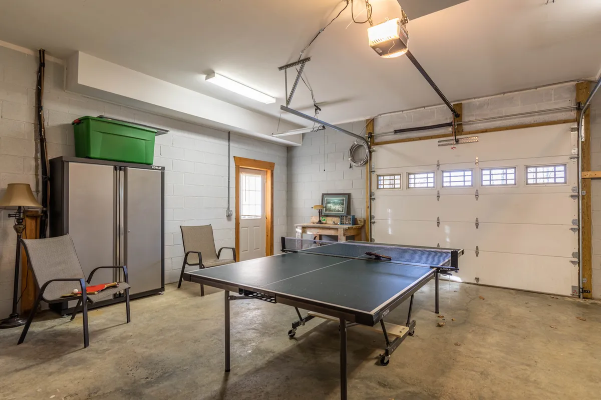 A bright, open area with a ping pong table as the centerpiece, garage doors letting in natural light, a few chairs for spectators or players, and a storage cabinet along the wall. The setup encourages fun group activities and relaxed gatherings.