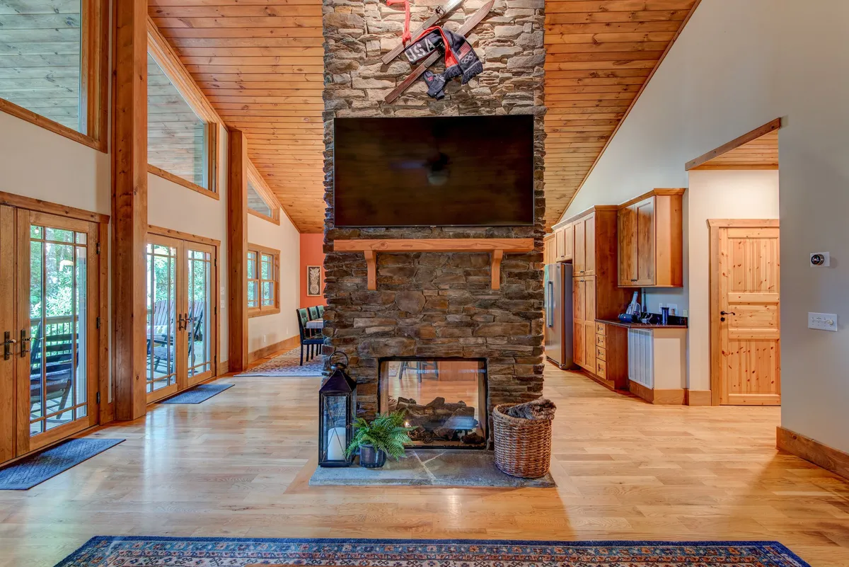 An open-concept living area showcasing a striking see-through stone fireplace, exposed wooden beams, large windows with forest views, and a glimpse of the kitchen area. The room is arranged for gatherings with ample comfortable seating and a warm, natural glow.