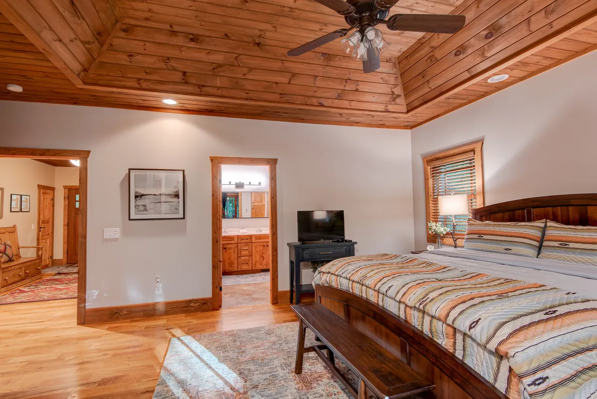 A bedroom with a serene, comfortable atmosphere, featuring a ceiling fan, striped bedding, a wooden bench at the foot of the bed, and natural light streaming in. The space is designed for relaxation, reading, or watching TV, aligning with the tranquil sanctuary theme.