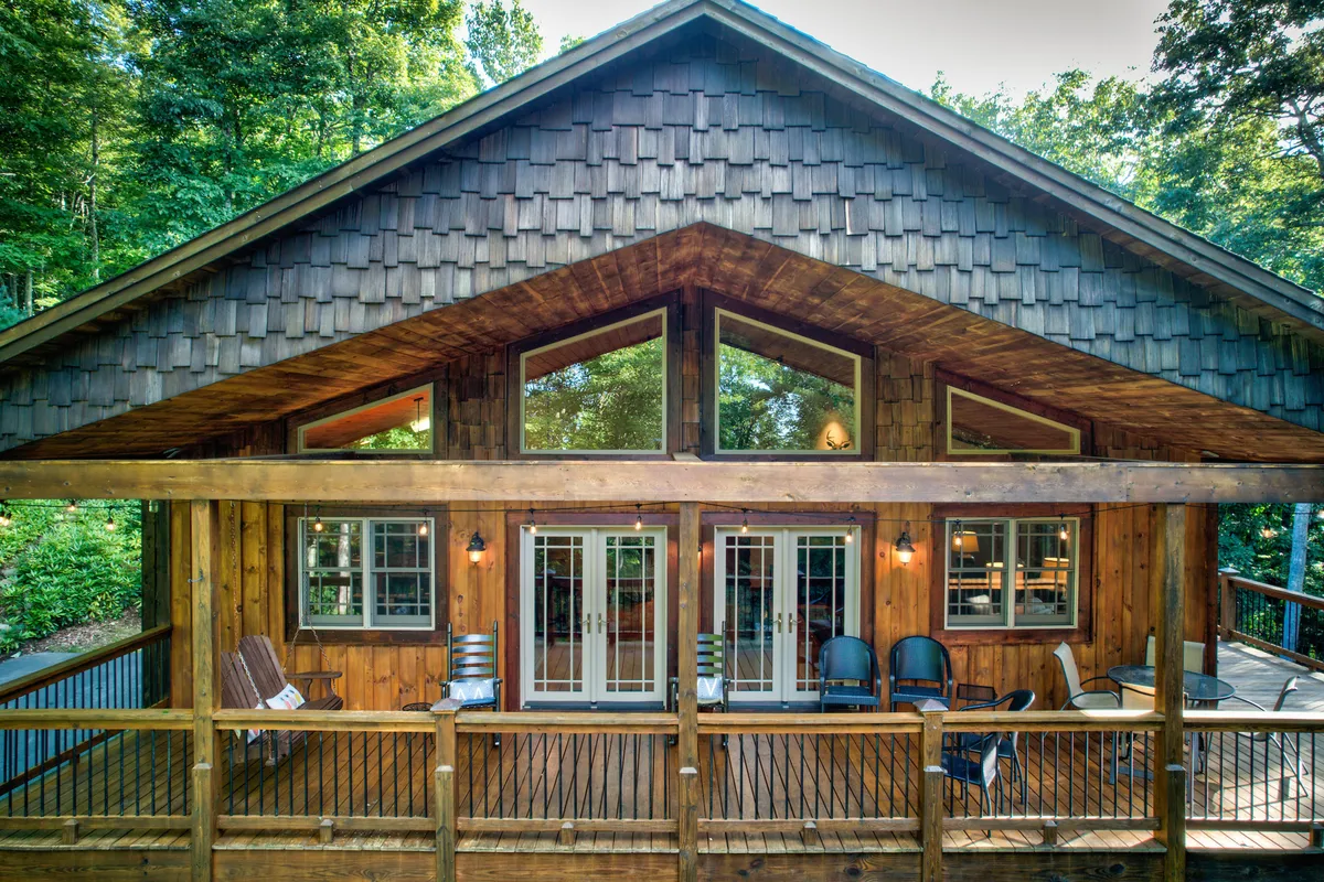 This image features a spacious wooden deck with comfortable seating, large windows that connect the indoors to the outdoors, and a shingle roof. The surrounding greenery provides a natural, serene backdrop, making the deck an ideal spot for gatherings or quiet relaxation.