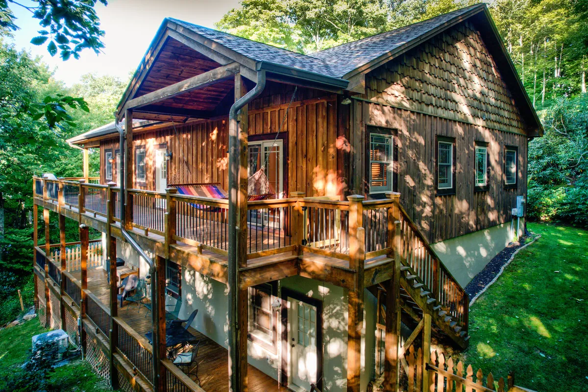 A spacious wooden deck with rustic charm, featuring large windows that reflect the serene, leafy surroundings. The deck is framed by lush greenery, offering a tranquil environment for relaxation, socializing, or stargazing. The wood siding and natural textures blend seamlessly with the landscape, reinforcing the retreat vibe.
